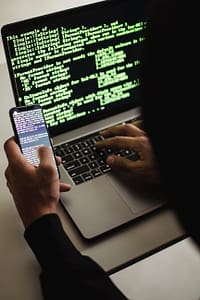 unrecognizable hacker with smartphone typing on laptop at desk